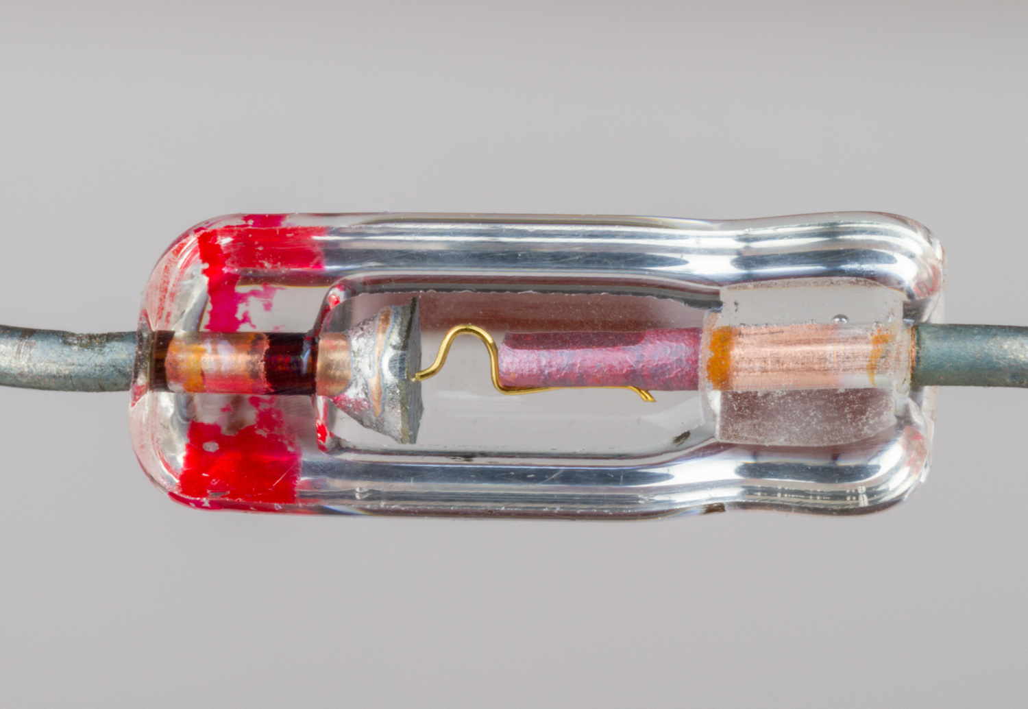 A closeup of a germanium 'cat whisker' diode, as one might find in a crystal radio. It is a clear glass horizontal tube, with splotches of red paint on the left hand side; in the center is a metal-colored cone touching a small copper-colored hook, which connects to a red stick